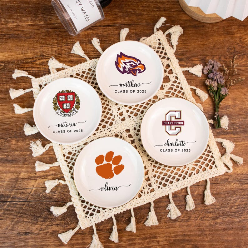 May include: Four white ceramic trinket dishes with gold rims. Each dish features a different design: a university crest, a paw print, and two university logos. Names and the text "Class of 2025" are also printed on the dishes.