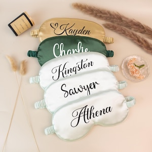 May include: A collection of personalized satin sleep masks in various colors, including gold, green, and white. Each mask features a name in elegant script. The names are Kayden, Charlie, Kingston, Sawyer, and Athena. A small candle and decorative elements are also present.