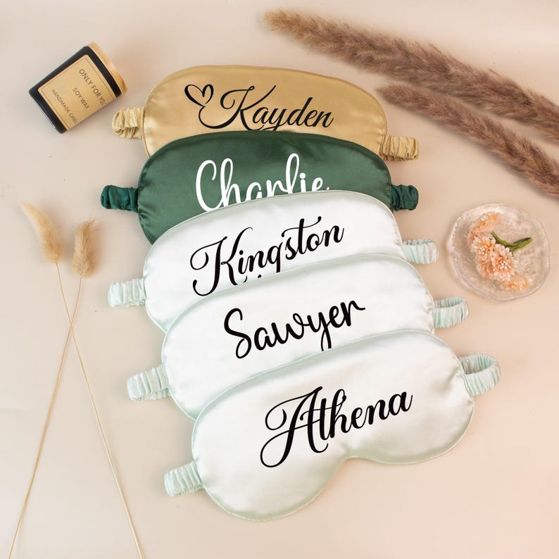 May include: A collection of personalized satin sleep masks in various colors, including gold, green, and white. Each mask features a name in elegant script. The names are Kayden, Charlie, Kingston, Sawyer, and Athena. A small candle and decorative elements are also present.