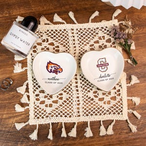 May include: Two heart-shaped ceramic trinket dishes with gold rims, personalized with names and graduation years. One features an eagle logo, the other a university logo. A perfume bottle, rings, and a decorative doily are also present.