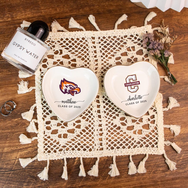 May include: Two heart-shaped ceramic trinket dishes with gold rims, personalized with names and graduation years. One features an eagle logo, the other a university logo. A perfume bottle, rings, and a decorative doily are also present.