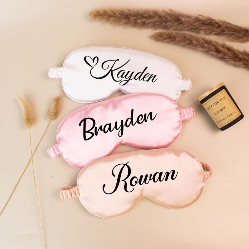 May include: Three satin sleep masks in white, pink, and peach, each with a name in black script: Kayden, Brayden, and Rowan. A small candle with text is visible. The masks have elastic bands and are designed for eye coverage.