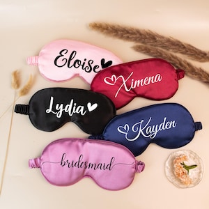 May include: Five satin sleep masks in pink, burgundy, black, navy blue, and lavender. Each mask has a name or word in white script. The pink mask says "Eloise", the burgundy mask says "Ximena", the black mask says "Lydia", the navy blue mask says "Kayden", and the lavender mask says "bridesmaid".