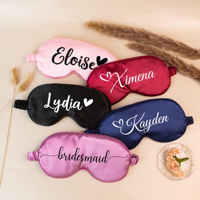 May include: Five satin sleep masks in pink, burgundy, black, navy blue, and lavender. Each mask has a name or word in white script. The pink mask says "Eloise", the burgundy mask says "Ximena", the black mask says "Lydia", the navy blue mask says "Kayden", and the lavender mask says "bridesmaid".