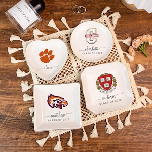 May include: A collection of white ceramic trinket dishes with gold rims. One is heart-shaped with a paw print and the name "Olivia." Others feature college logos and names like "Charleston," "Matthew," and "Victoria," with "Class of 2025."
