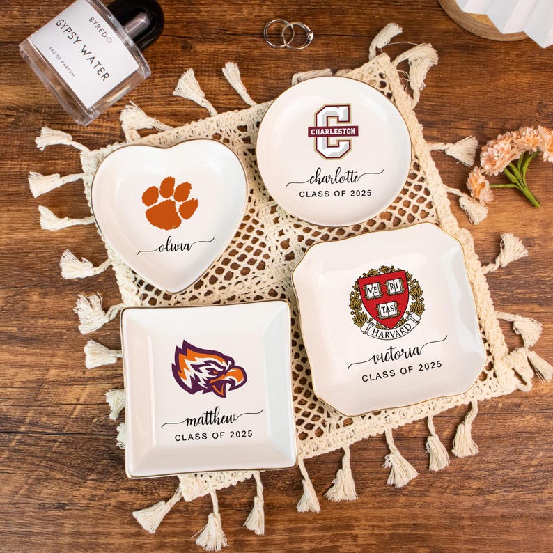 May include: A collection of white ceramic trinket dishes with gold rims. One is heart-shaped with a paw print and the name "Olivia." Others feature college logos and names like "Charleston," "Matthew," and "Victoria," with "Class of 2025."