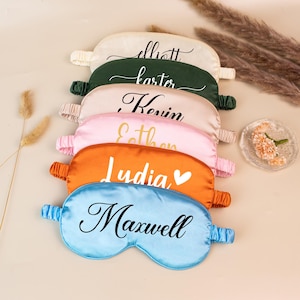 May include: A stack of personalized sleep masks in various colors, including cream, green, pink, orange, and blue. Each mask features a name in elegant script. The orange mask has a heart symbol next to the name.