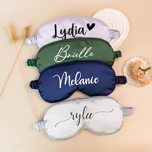 May include: Four satin sleep masks in lavender, forest green, navy blue, and silver. Each mask has a name printed in white script: Lydia, Brielle, Melanie, and Rylee. The masks have elastic bands and are arranged on a light beige surface.