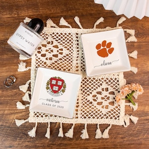 May include: A flat lay featuring decorative trinket dishes, perfume, and rings on a cream-colored, tasseled doily. Two white square dishes have gold trim and personalized text. One dish has a paw print, the other has a Harvard crest.