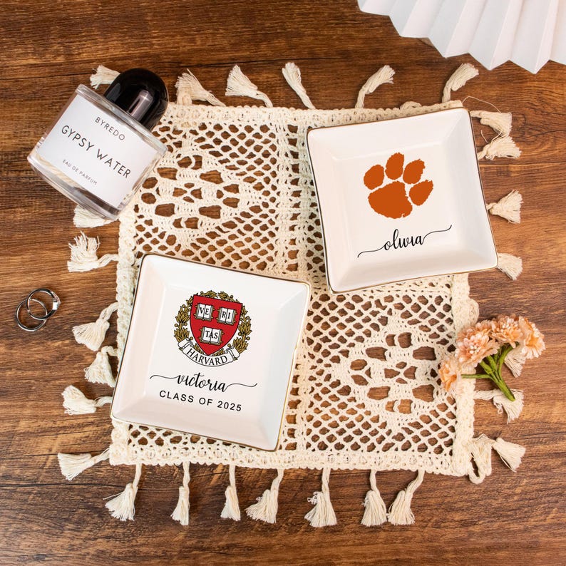 May include: A flat lay featuring decorative trinket dishes, perfume, and rings on a cream-colored, tasseled doily. Two white square dishes have gold trim and personalized text. One dish has a paw print, the other has a Harvard crest.