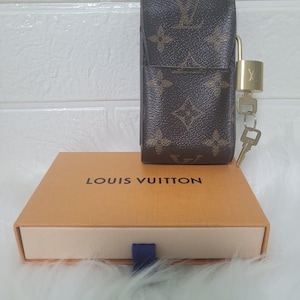 Puede incluir: Un estuche marrón y beige con monograma y un candado y llave dorados, apoyado sobre una caja beige con la inscripción "LOUIS VUITTON". El estuche tiene forma rectangular y cierre de solapa. La caja es rectangular con interior blanco.