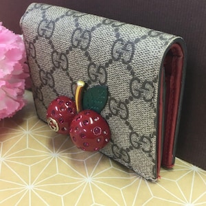 May include: A brown and beige wallet with a repeating logo pattern. The wallet features a red interior and a decorative cherry embellishment with red gems and a gold stem. The cherry is attached to the front of the wallet.