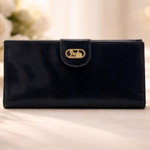 Puede incluir: Una cartera de cuero azul marino con un cierre dorado con un diseño de carruaje tirado por caballos. La cartera rectangular está cerrada, con un acabado liso y brillante. Se ven rosas blancas en el fondo.