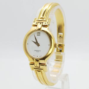 Puede incluir: Un reloj de pulsera Givenchy Paris dorado con esfera blanca y agujas negras. El reloj tiene una correa y un cierre dorados. La esfera del reloj tiene el logotipo de Givenchy y las palabras "SWISS MADE".