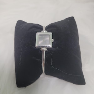 Puede incluir: Un reloj de pulsera plateado con una esfera cuadrada y una banda delgada. El reloj se muestra sobre un cojín de terciopelo negro sobre un fondo blanco y esponjoso. La esfera del reloj tiene un diseño simple y minimalista.