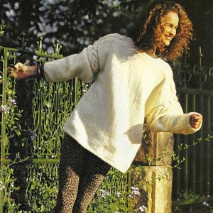May include: A cream-coloured jumper with cable trim is worn with leopard print leggings and black boots. The jumper has a relaxed fit. The text "Lady's jumper with cable trim" is visible. The image is set outdoors with a green gate and stone wall.