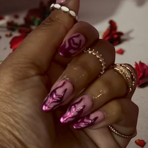 Puede incluir: Una mano con uñas largas y rosas con un diseño de llama magenta metálico. Las uñas están adornadas con anillos, incluido un anillo de cuentas blancas y anillos dorados. Pétalos de rosa rojos están esparcidos en el fondo.
