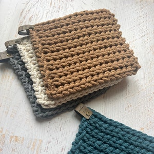 May include: Four crocheted pot holders in different colors: brown, white, gray, and teal. Each pot holder has a leather tag with the word "handmade" written on it.