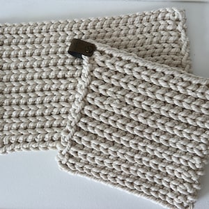 May include: Two off-white, rectangular woven placemats with a thick, textured rope-like design. Each placemat has a small, rectangular, dark metal accent piece attached to one corner. The placemats are a neutral color.