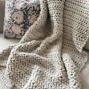 May include: A chunky, hand-crocheted throw blanket in a solid light beige color. The blanket features a thick, textured yarn construction with visible interlocking loops and a dense, heavy weave that drapes over the surface of a neutral-toned sofa.