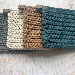 May include: Four handmade crochet pouches in shades of gray, cream, brown, and teal. Each pouch features a textured, knotted stitch design and a leather tag with a metal rivet. The pouches are arranged side by side.