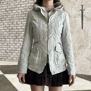 May include: A pale grey hooded jacket with a zip, pockets, and a textured finish. The jacket is worn over a white top and a dark brown skirt. The background includes a brick wall and a decorative sword.