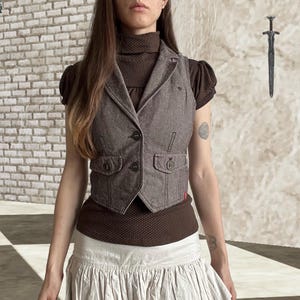 May include: A brown vest with a notched lapel, two front pockets with button closures, and a two-button front fastening. It is worn over a brown short-sleeved top featuring a small white polka dot pattern and a high turtleneck collar.