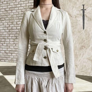 May include: A light beige blazer with a belted waist and four button closure. The blazer has two front pockets and a notched collar. The model is wearing a black top and a light beige skirt.