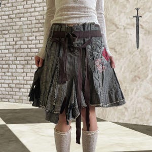 Puede incluir: Falda a rayas grises y marrones con cinturón y lazo marrón. La falda tiene un aspecto desgastado con bordes deshilachados y parches de tela. Un parche floral es visible en el lado derecho. La falda es hasta la rodilla.