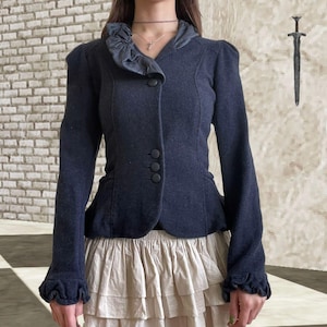 Peut inclure: Veste bleu marine foncé avec un col et des poignets à volants. La veste a trois boutons et une silhouette ajustée. Les manches ont un léger gonflement aux épaules. La modèle porte une jupe beige.