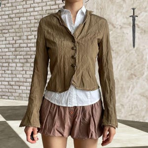 Può includere: Giacca marrone strutturata con quattro bottoni, indossata sopra una camicia bianca con colletto e abbinata a pantaloncini rosa. La giacca ha maniche lunghe e un aspetto stropicciato unico. I pantaloncini hanno una chiusura con bottoni.