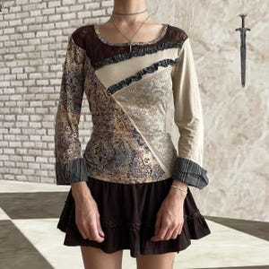 Op de afbeelding: Een top met lange mouwen met een patchwork-ontwerp met paisley- en effen stoffen in bruin, beige en patronen. De top heeft een vierkante halslijn en een ruchesrand. De manchetten hebben een ruitpatroon. Een bruine rok met een geplooid zoom maakt de outfit compleet.