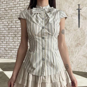 Puede incluir: Una blusa a rayas con un lazo en el cuello. La blusa presenta rayas verticales en tonos grises, blancos y un toque de verde. La prenda tiene mangas cortas y se combina con una falda beige escalonada, creando un conjunto de inspiración vintage.