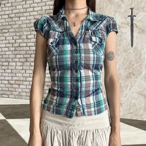 May include: A short-sleeved button-down shirt featuring a plaid pattern in shades of green, navy blue, and white. The shirt includes a classic collar, two chest pockets with flaps, and a row of dark buttons running down the centre front.