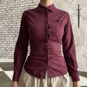 May include: A burgundy button-down shirt with a small, repeating dot pattern. The long-sleeved shirt has a fitted silhouette, a chest pocket, and a classic collar. The shirt is paired with a light-colored skirt.