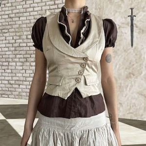 May include: A beige vest with a notched collar and four buttons over a brown blouse with short, puffed sleeves. The blouse has a ruffled neckline with white trim. The outfit is paired with a light-colored skirt.