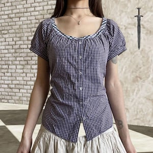 May include: A purple and white checkered blouse with short sleeves and a scoop neckline. The blouse has a button-up front and a contrasting green and white trim along the neckline. The fabric appears to be lightweight, suitable for warmer weather.