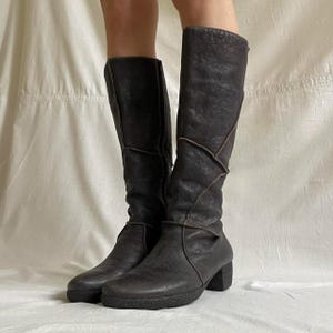 Puede incluir: Un par de botas altas de color marrón oscuro. Las botas tienen una apariencia de cuero texturizado con un tacón de bloque y una punta redondeada. El diseño incluye paneles cosidos y un sutil ribete en contraste. Las botas se muestran sobre un fondo neutro.