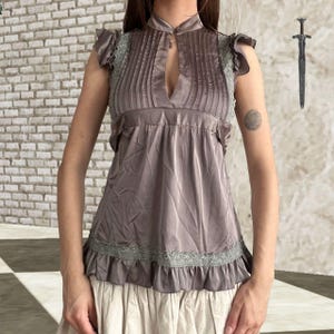 Pode incluir: Uma blusa sem mangas na cor taupe com gola alta e abertura central em forma de gota, pregas verticais no corpete e mangas com babados. A peça inclui acabamento em renda cinza claro ao longo dos ombros e na bainha com babados.