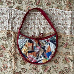 Może przedstawiać: Torba na ramię z patchworku z czerwonym wykończeniem i paskiem. Torba posiada różnorodne wzory i kolory tkanin, w tym niebieski, pomarańczowy i biały. Czerwona kokarda jest zawiązana na górze torby.