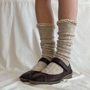 Op de afbeelding: Bruine leren Mary Jane-schoenen met een bandje en gesp, gedragen met beige slouch sokken met een kanten rand. De schoenen hebben een ronde neus en sierstiksels. De sokken zijn lichtbeige.