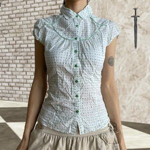 May include: White short-sleeved blouse with a small blue floral pattern and green trim. The blouse has a button-down front with green buttons and a rounded collar. The style is vintage and feminine.