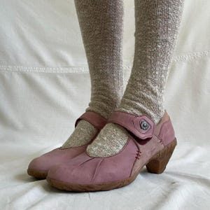 May include: A pair of dusty rose Mary Jane style shoes with a strap closure and a brown heel. The shoes have a textured appearance and are worn with gray socks.