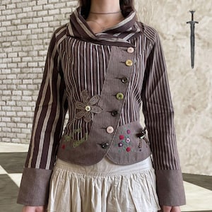 Könnte beinhalten: Braune gestreifte Jacke mit einem einzigartigen asymmetrischen Design. Sie hat einen hohen Kragen, mehrere Knöpfe und Blumenstickereien. Die Jacke hat ein Patchwork-Design mit einer Blumenapplikation und dem Text "BEHIND!" in Grün.