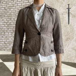May include: A brown blazer with three-quarter sleeves, a notched collar, and button details. The blazer is worn over a light blue blouse and a beige skirt. A decorative sword is in the background.