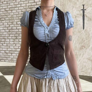 Puede incluir: Un chaleco de ante marrón con cierre de un solo botón sobre una blusa azul claro con mangas con volantes. La blusa se combina con una falda beige. Una ilustración de espada plateada está en el fondo.