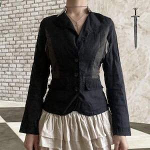 Puede incluir: Un blazer negro de manga larga con cuello de solapa y tres botones frontales, con paneles laterales en color oliva oscuro y dos bolsillos con solapa. El blazer se lleva sobre una falda beige de volantes superpuestos. Se ve un icono de espada plateada.