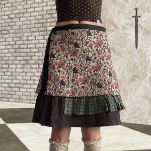 Puede incluir: Falda a capas con un estampado floral en rosa y morado sobre fondo crema. La falda tiene capas verdes oscuras y marrones. Prenda de vestir única.