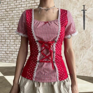 May include: A red and white patterned top with a square neckline and short sleeves. The top features a gingham pattern on the bodice and a floral print on the sides. White lace trim and a red ribbon corset detail add a decorative touch. The top is a vintage style.
