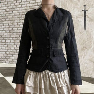 May include: Black blazer with contrasting panels on the upper chest and back. The blazer features a notched lapel, three buttons, and two pockets. The sleeves are long, and it is paired with a tiered, cream-coloured skirt.